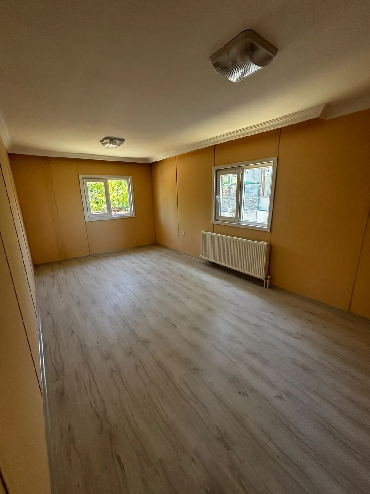 İki PVC pencereli, radyatörlü ve açık renk laminat parkeli prefabrik ev iç mekanı